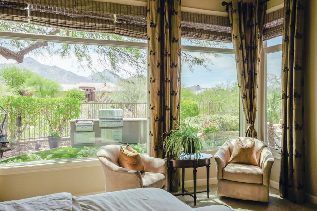 Sunlit interior sitting area with two upholstered armchairs and a small round table by large windows, looking out onto a landscaped backyard with a built-in outdoor grill, desert plants, and distant mountains framed by patterned curtains.