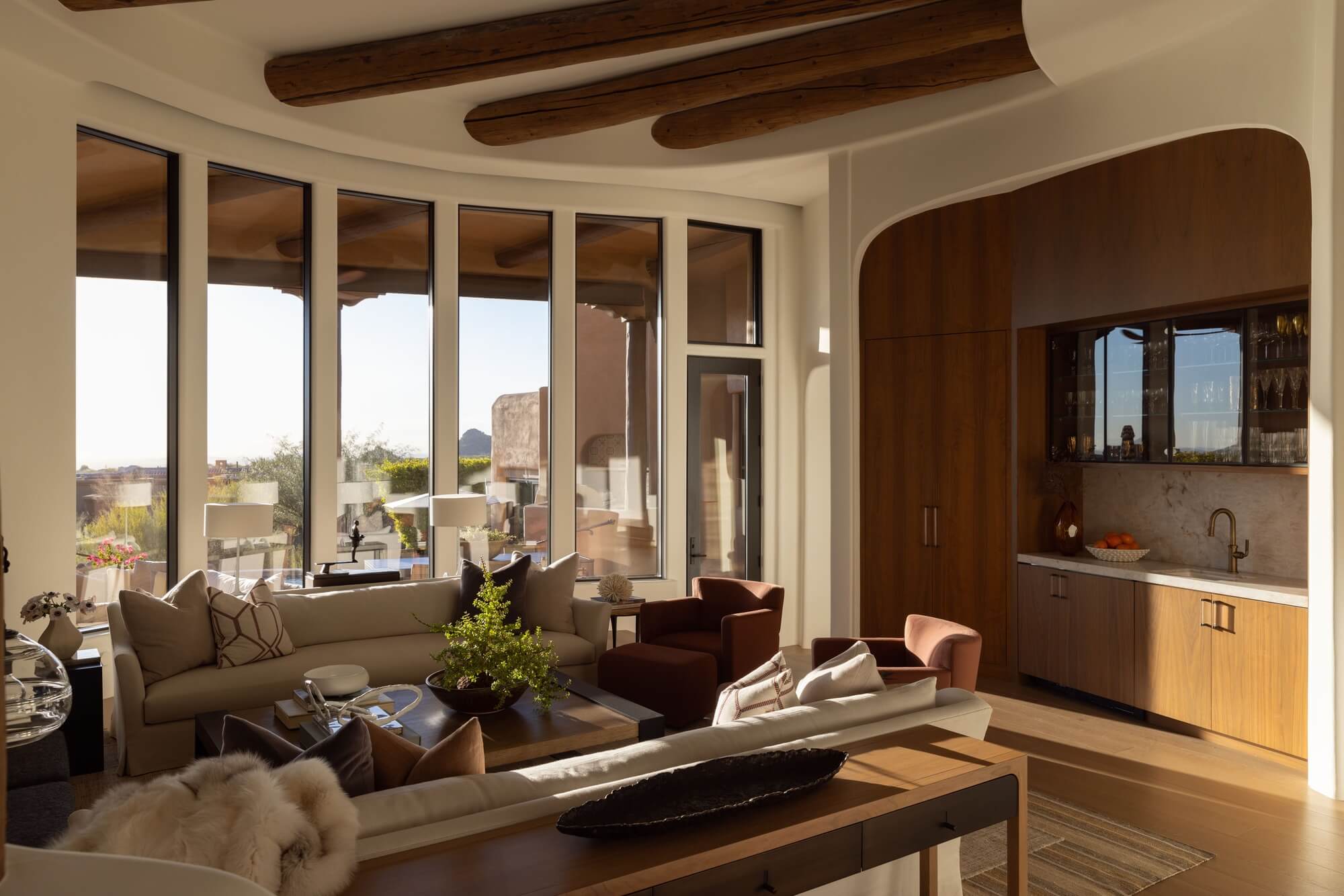 Sunlit living room with floor-to-ceiling windows, wood ceiling beams, neutral seating, and a built-in wood cabinet with a small bar area.
