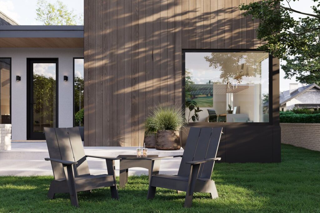 Modern home exterior with a wood-paneled accent wall and large black-framed window, overlooking a small patio setup with two dark Adirondack chairs and a matching table on green grass. Sunlight casts tree shadows across the facade, and a large potted plant and indoor dining area are visible through the window.