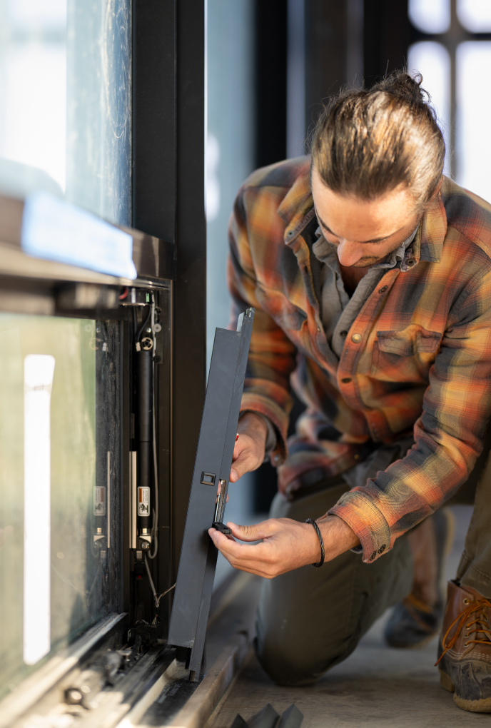 A Mountain West Windows and Doors technician kneels indoors while repairing or adjusting a window frame using hand tools.